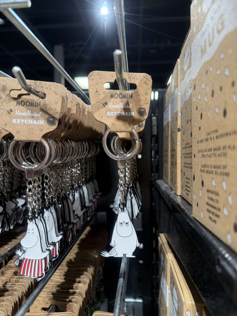 A close-up of Moomin keychains lined up on a store display rack. The focus is on a single keychain in the center featuring a Moomin with a grumpy expression.