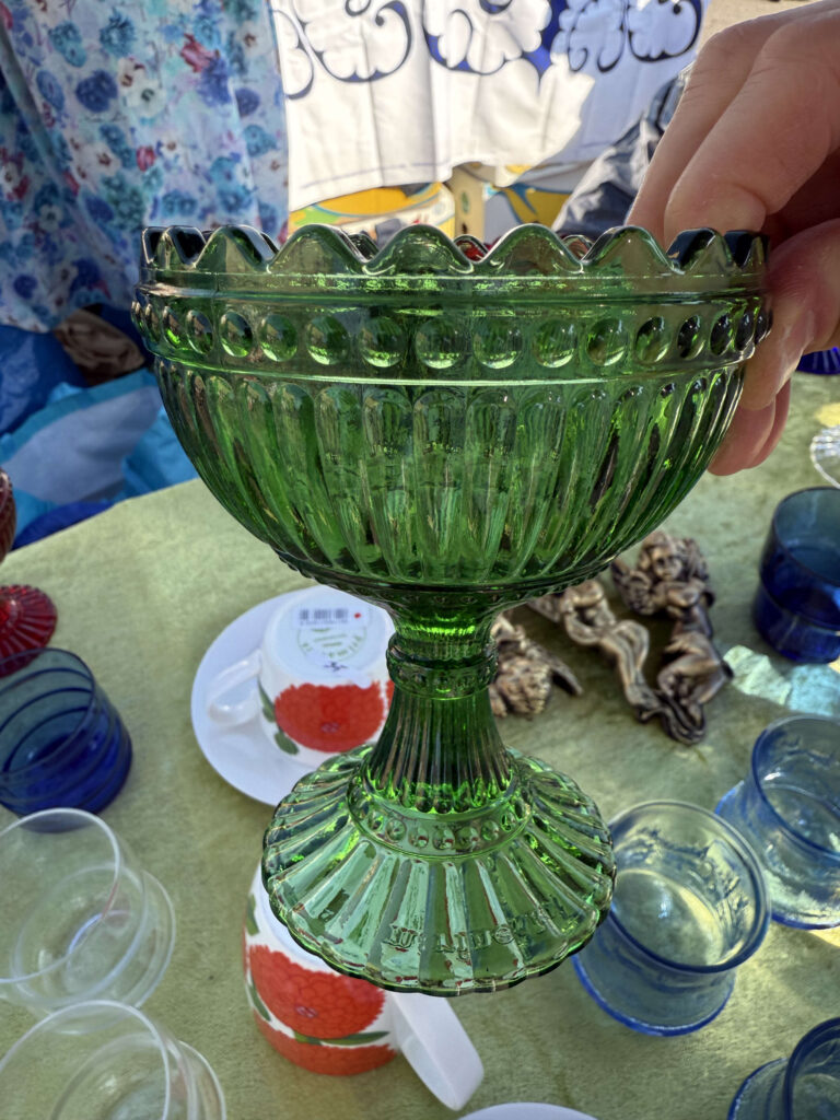 A hand holding a green glass Maribowl at a flea market. The bowl is shining in the sunlight, and a table with other tableware is visible in the background.