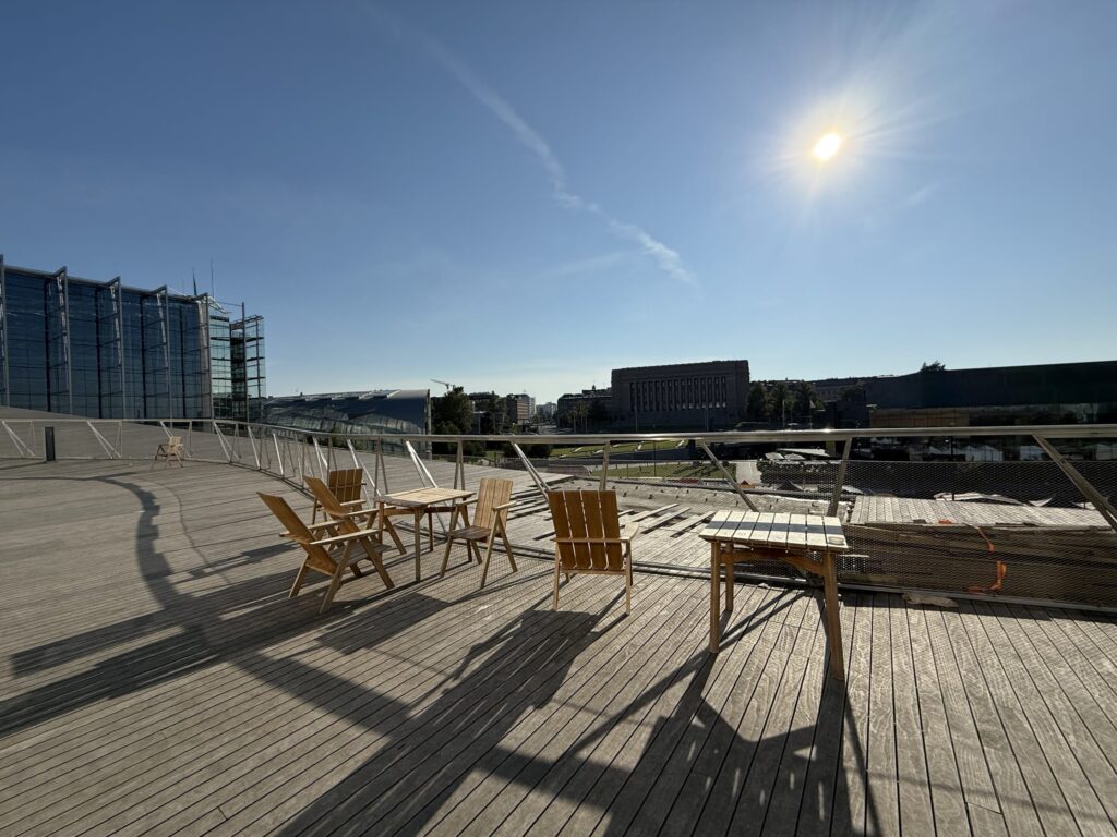 晴れた日のウッドデッキのテラス。木製のテーブルと椅子が置かれ、背景にはヘルシンキの近代的な建物と青空が広がっている。太陽が強く照りつけ、長い影ができている。