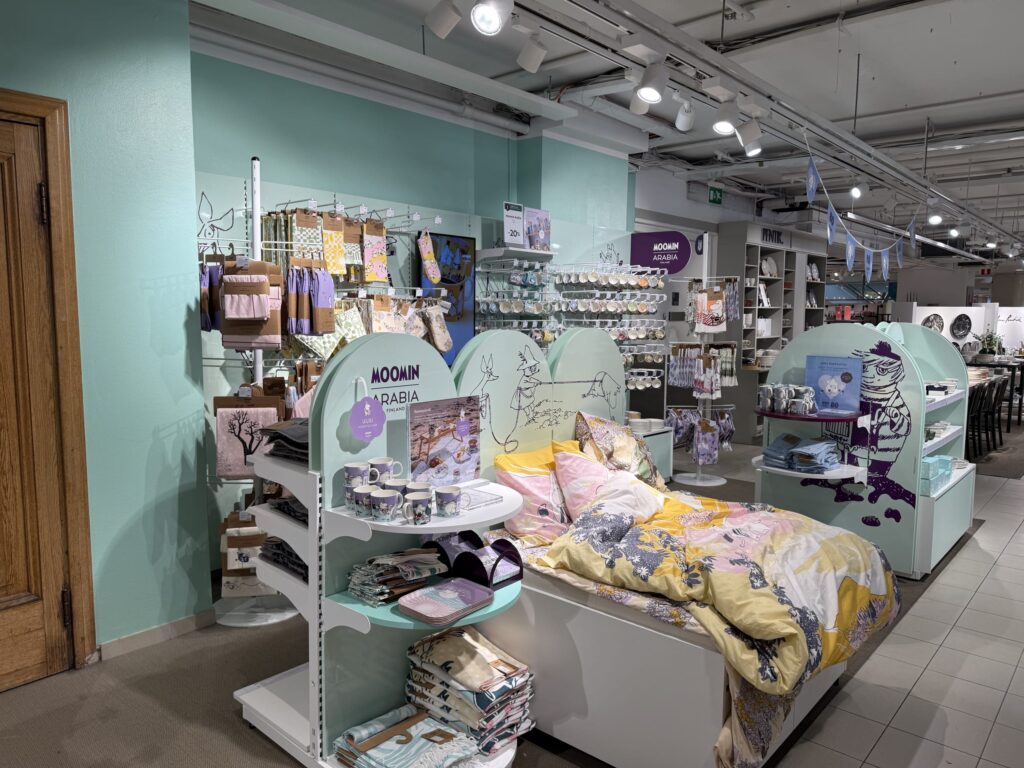 The Moomin by Arabia home goods section in a department store. Mint green shelves are filled with mugs and towels, and a bed with Moomin-patterned linens is set up in the center.