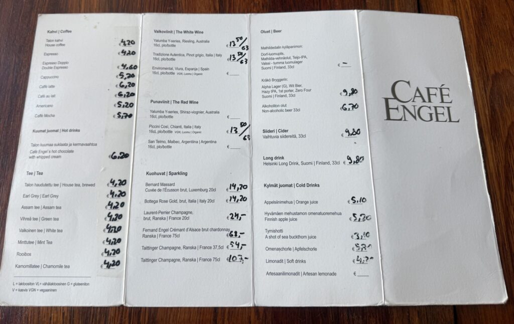 A drink menu from Café Engel in Helsinki is spread open on a dark wooden table, showing categories for coffee, wine, and beer in Finnish and English.