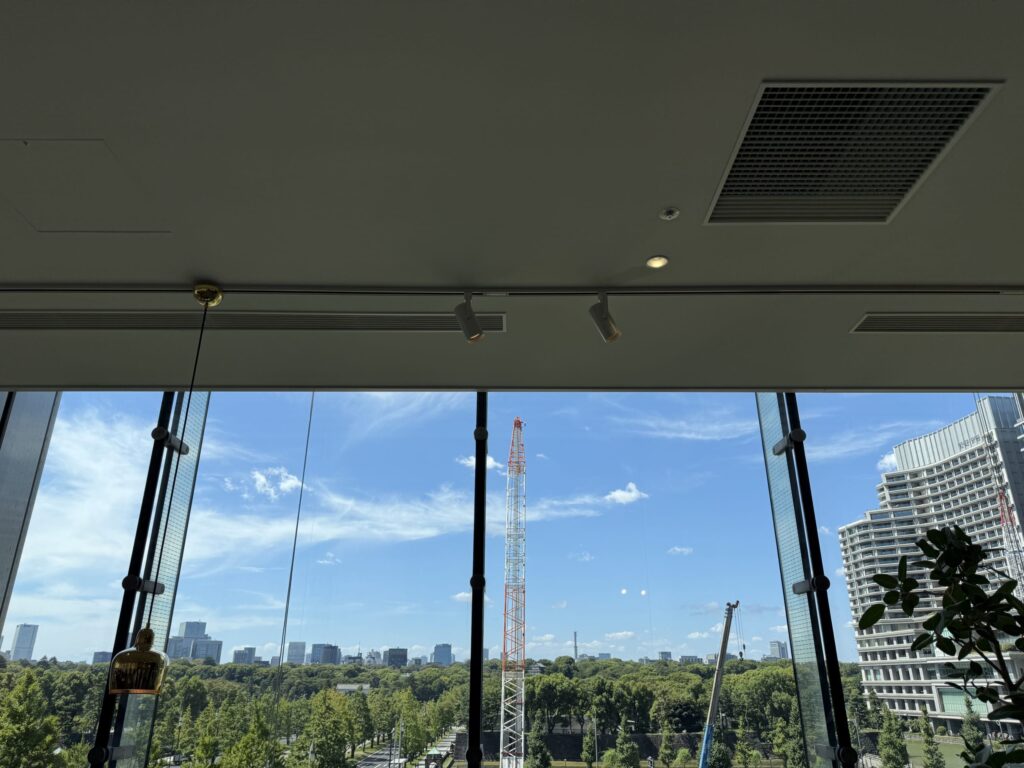 A view of a city landscape from a large window. A lush green park spreads out in the foreground, with a skyline of buildings in the distance. A construction crane stands tall against the blue sky.