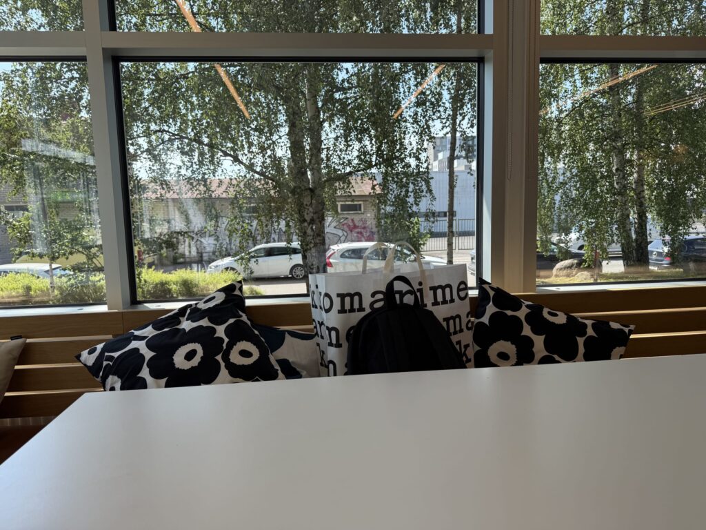A sofa seat by a large window, featuring Marimekko cushions and a logo bag, with a view of birch trees and parked cars outside.