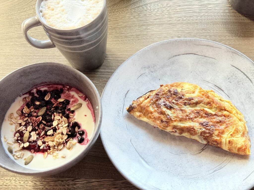 A breakfast scene on a wooden table, featuring a plate with an omelette, a bowl of yogurt with granola and berry sauce, and a mug of frothed hot latte.