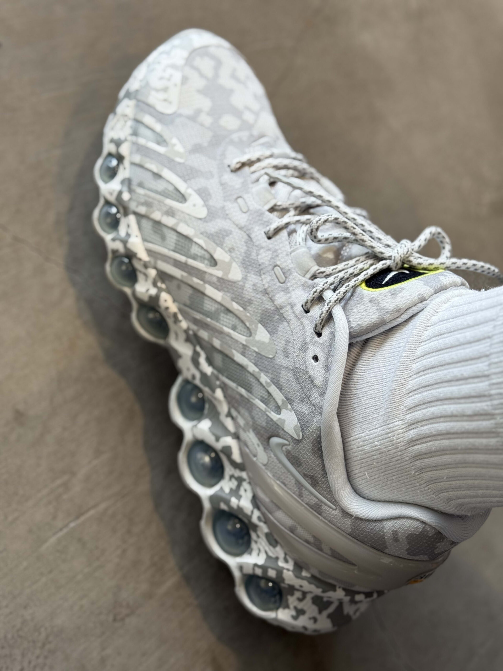 グレーと白のデジタルカモフラージュ柄が施されたC.E. x Nike Air Max 98 SPスニーカーを履いた足元のクローズアップ。特徴的なバブル状のソールユニットが見える。