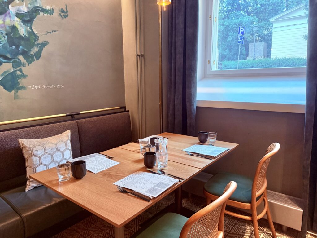A wooden table set with menus at a restaurant, located by a window. There is a booth seat on one side and rattan chairs on the other.