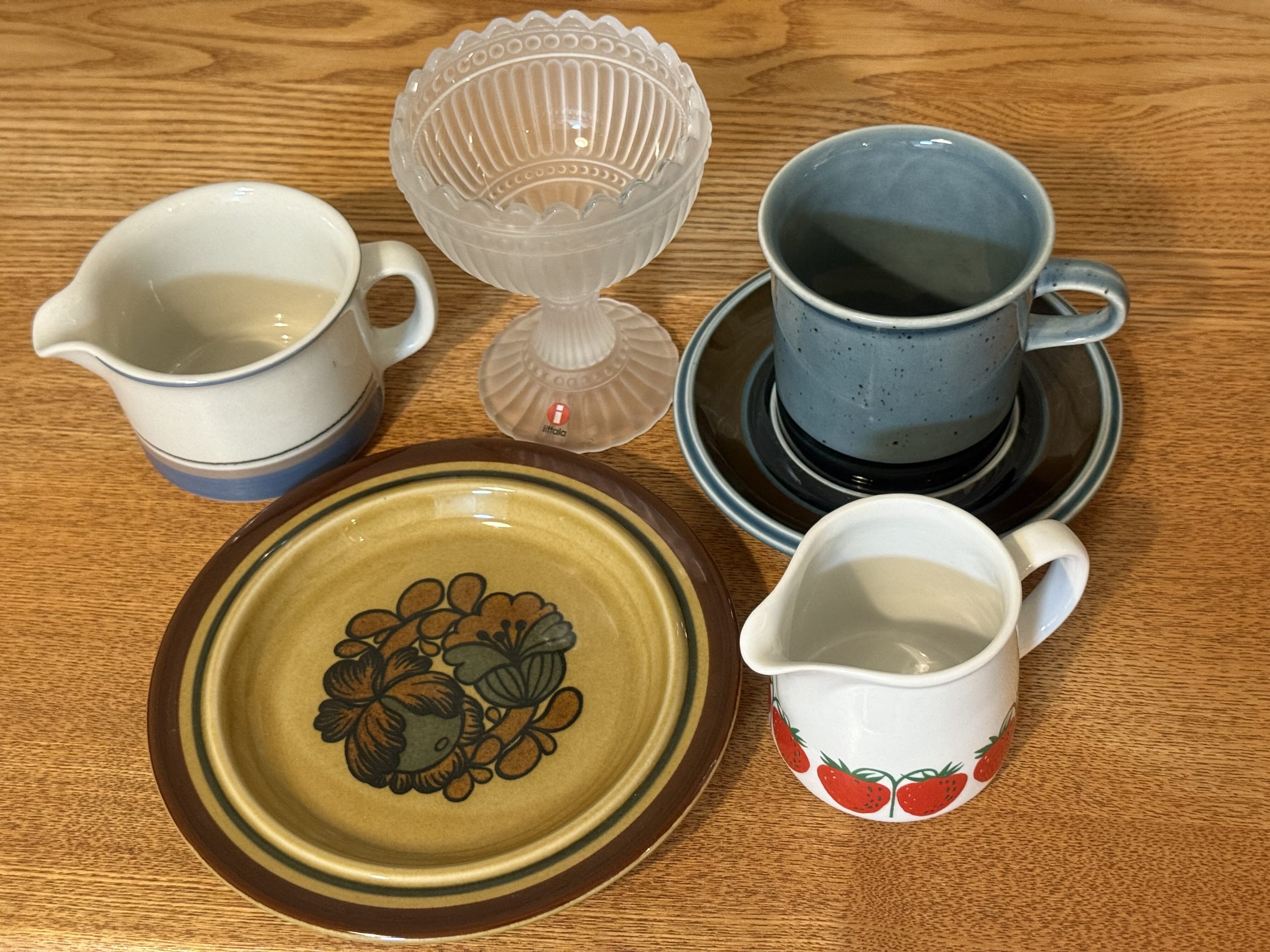 Five pieces of vintage tableware on a wooden table: a blue cup and saucer, a yellow plate with a floral pattern, a creamer with stripes, a creamer with a strawberry pattern, and an iittala glass dessert bowl.