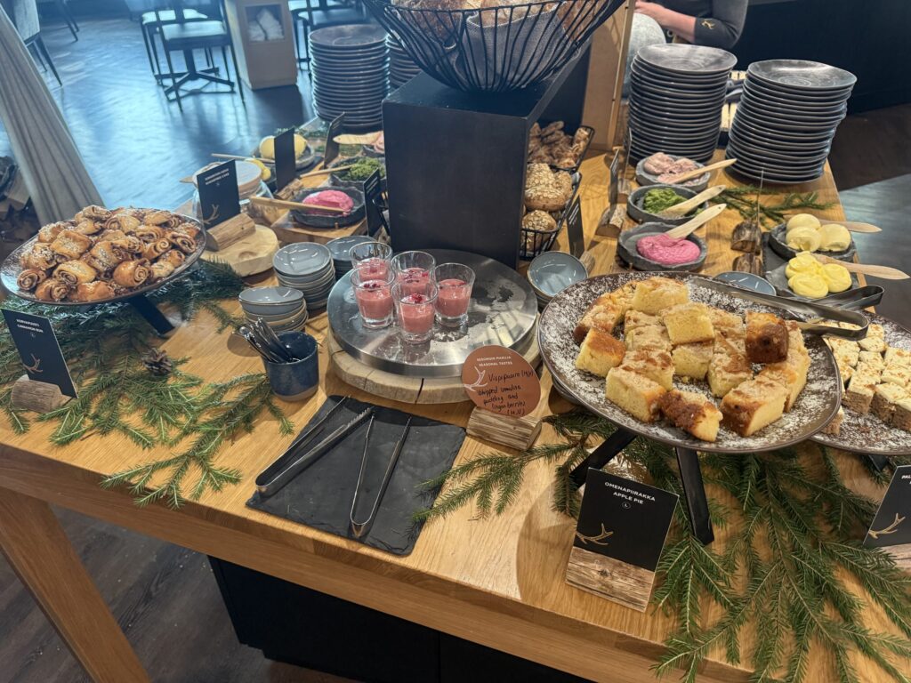 A buffet spread on a wooden table. It features cinnamon rolls, cakes, a pink pudding, various breads, and is decorated with pine branches.