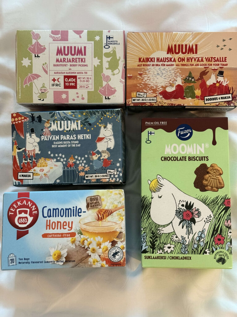 Five boxes of tea and biscuits on a white surface: three boxes of Moomin-themed tea, one box of Moomin chocolate biscuits by Fazer, and one box of Teekanne Camomile-Honey tea.