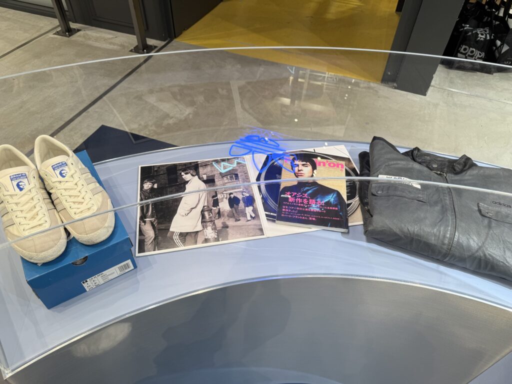 A display case with Adidas and Liam Gallagher collaboration items. It features a pair of cream-colored sneakers, a black and white photo of Oasis, a Japanese music magazine, and a black leather jacket.