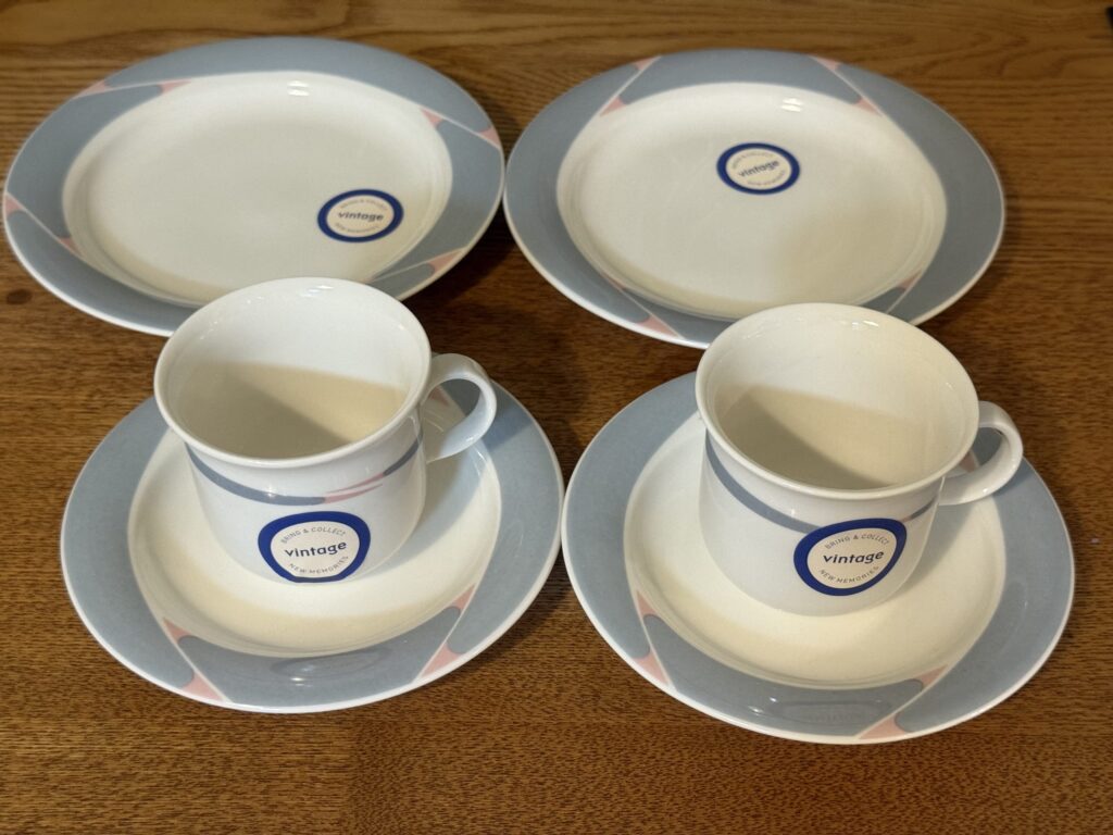 A vintage-style set of two teacups, two saucers, and two small plates with a geometric pattern in light blue and pink, arranged on a wooden table.
