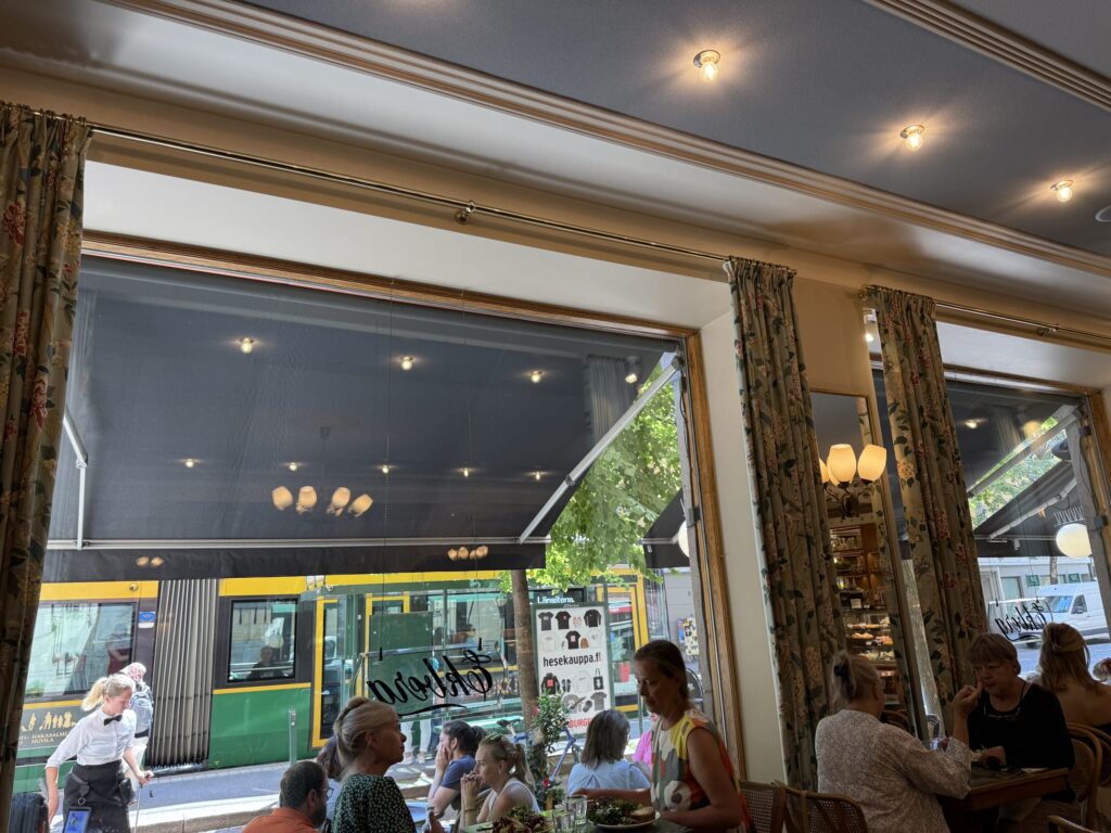 A view from a window seat in a classic cafe, looking out at a green and yellow tram stopped on the street. Inside, people are enjoying their meals and conversations.
