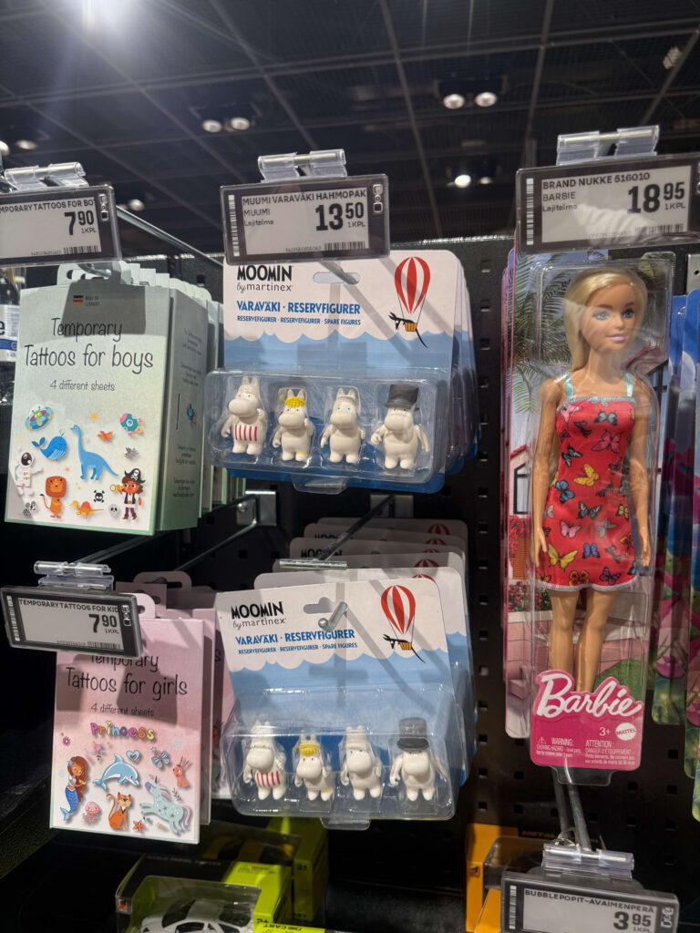 スーパーマーケットのおもちゃ売り場の棚に、ムーミンのフィギュアセット、バービー人形、子供向けのテンポラリータトゥーが並んでいる様子。
