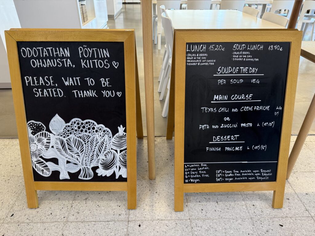 Two wooden-framed chalkboard signs in a modern cafe. The left board has handwritten seating instructions in Finnish and English, with white floral illustrations at the bottom. The right board displays a handwritten lunch menu including soup, main courses, and dessert.