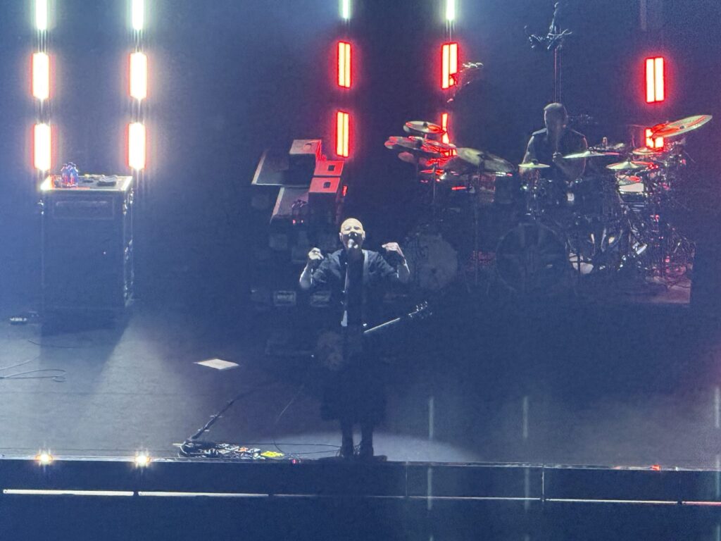 On a dark, smoky stage, a bald vocalist stands center stage with his arms outstretched towards the microphone. A drummer and vertical lights glowing red and white are visible in the background.