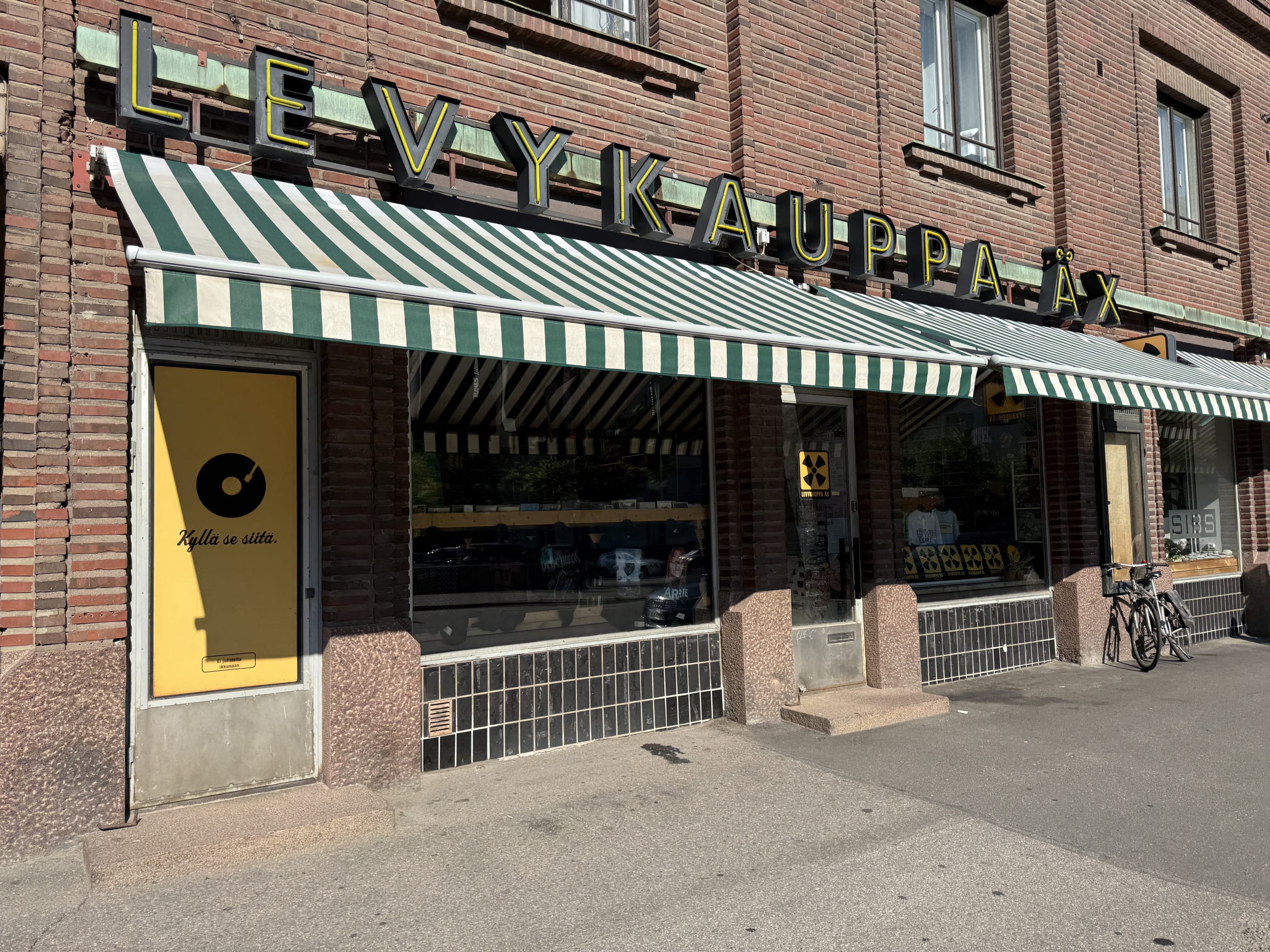 The storefront of the record shop 'Levykauppa Äx' in front of a brick building on a sunny day. It has large windows under a green and white striped awning, and a yellow neon sign displays the store's name.
