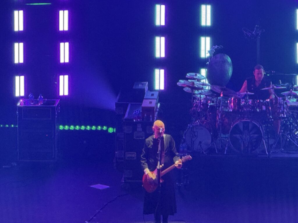 A concert scene showing Billy Corgan playing guitar and singing on a stage lit with purple lights, with the drummer performing behind him.