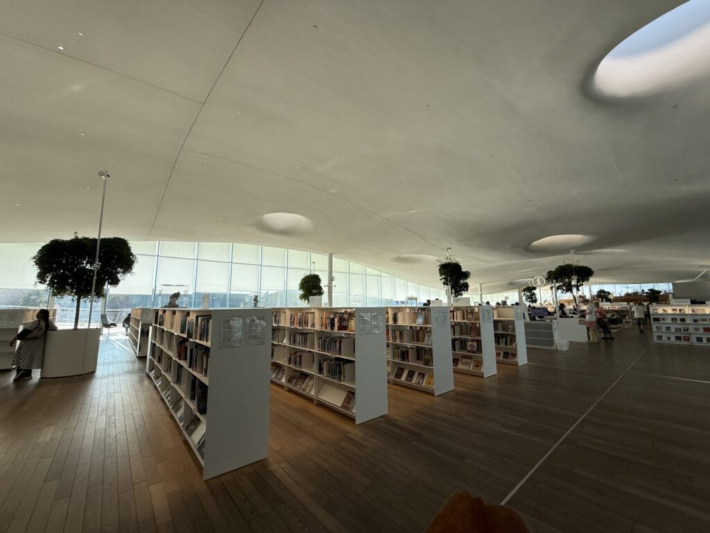 モダンな図書館の広々とした内部。波打つ白い天井の下に低い本棚が並び、人々が読書などを楽しんでいる。