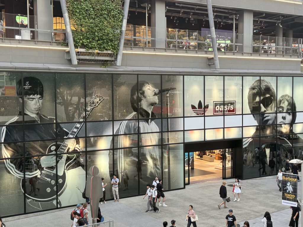 The exterior of the Adidas store at MIYASHITA PARK in Shibuya. The entire glass facade of the building is wrapped in giant black-and-white photographs of the Gallagher brothers from the British rock band Oasis. Many people are walking past the front of the store.