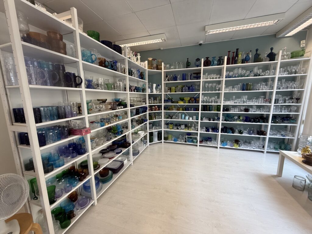 イッタラのカラフルなガラス製品が陳列された白い棚が並ぶ店内の様子。青、緑、琥珀色など様々な色のグラスや食器が棚を埋め尽くしている。