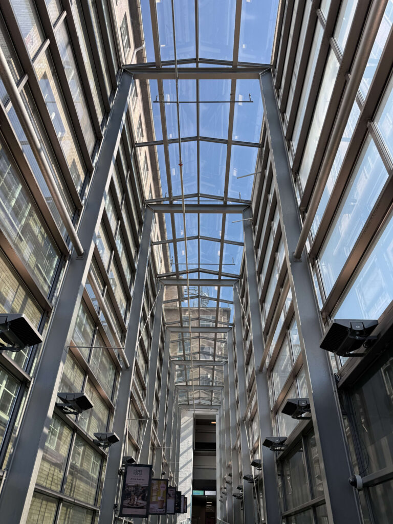 A low-angle view looking up through a modern corridor with glass walls and a glass ceiling supported by gray steel beams, revealing the blue sky above.