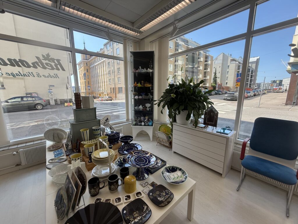 The interior of a vintage shop with bright light streaming through large windows. Various colorful vintage dishes, in blues and yellows, are displayed on a white table, with a view of a sunny city street outside.