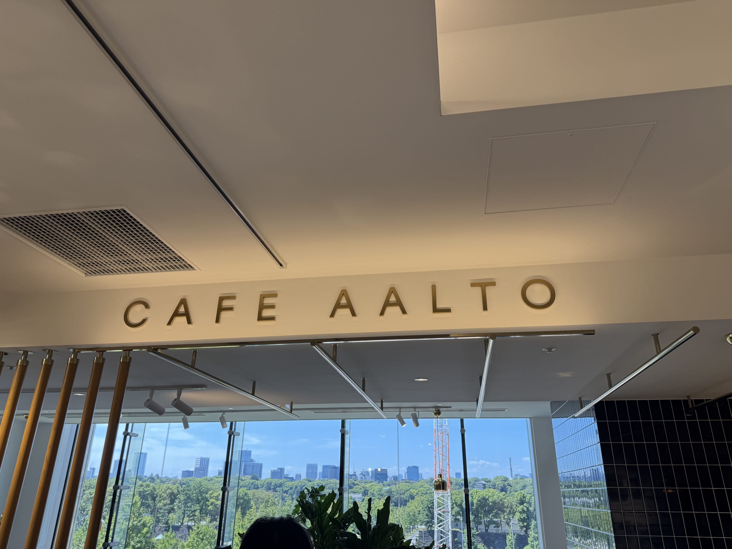 The interior of CAFE AALTO. The golden letters of 'CAFE AALTO' are on a white beam near the ceiling, and below it, a large window shows a view of a lush green park and city buildings.