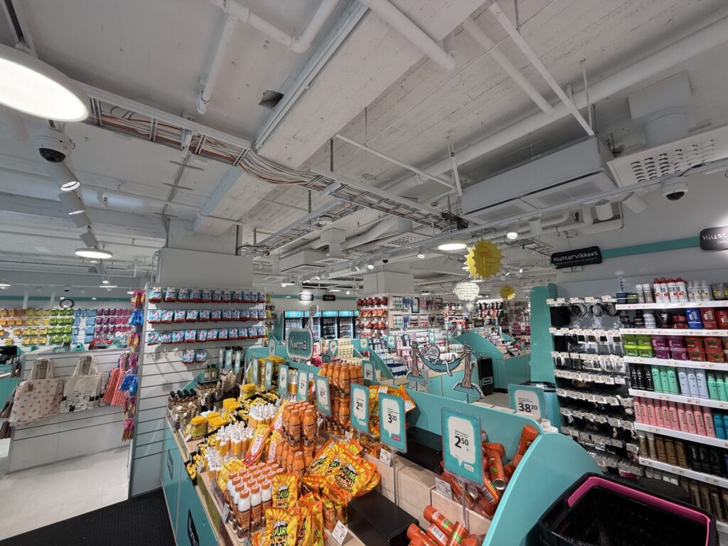 The interior of a brightly lit variety store with an exposed industrial ceiling, showing aisles stocked with snacks and toiletries.
