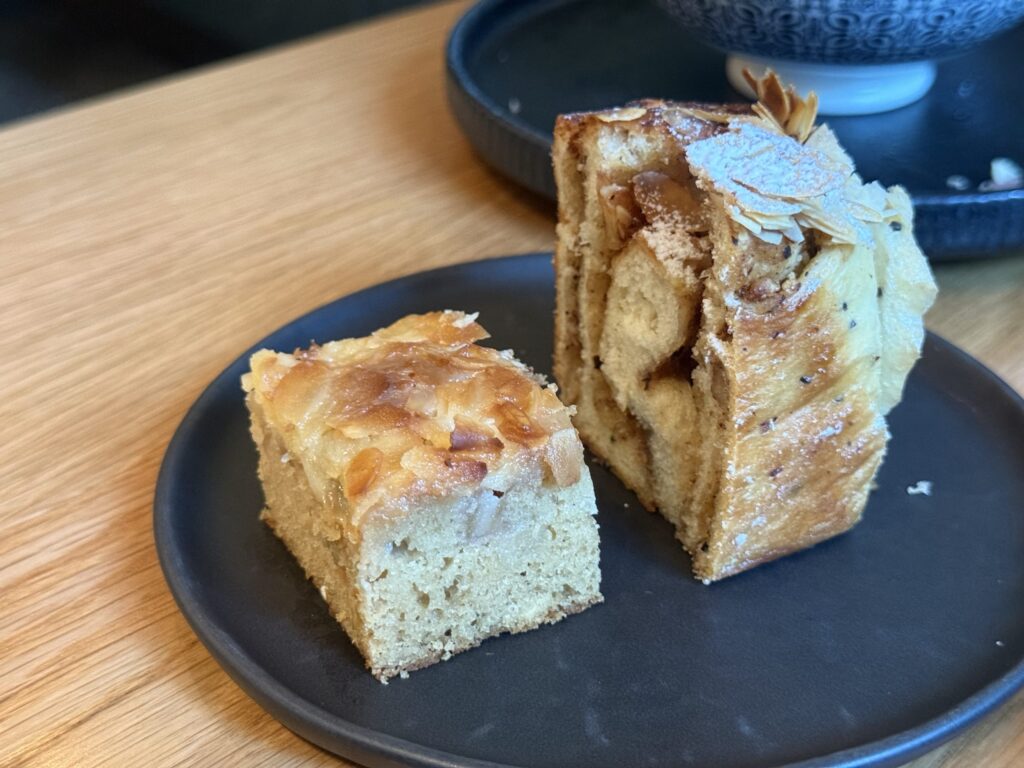 Two types of almond bread on a black plate on a wooden table. In the foreground is a square almond cake, and in the background is a layered bread with sliced almonds and powdered sugar.