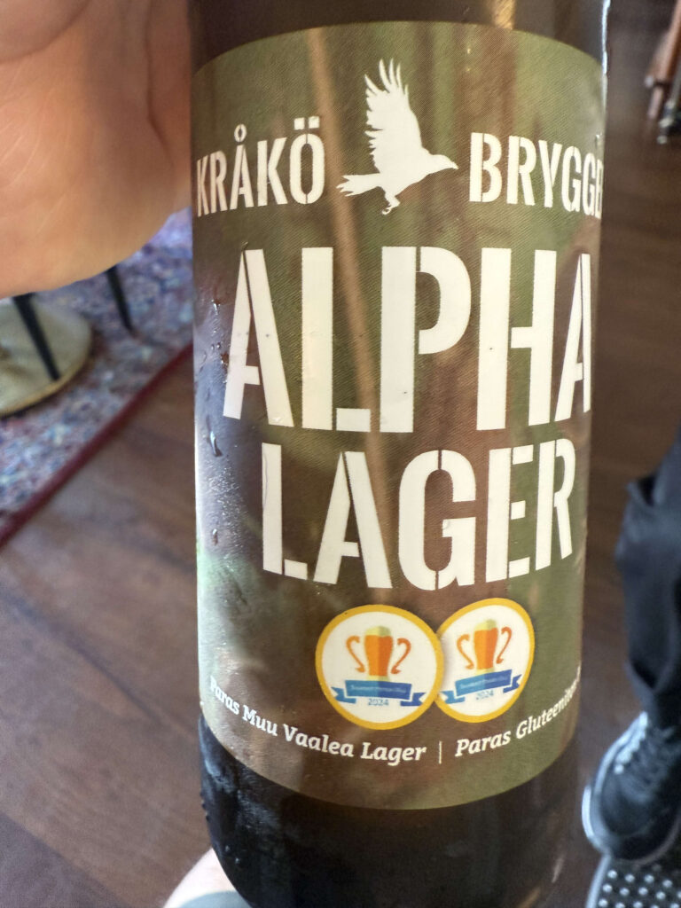 A hand holding a bottle of Kråkö Bryggeri's Alpha Lager. The green label features the product name in white stencil lettering, a silhouette of a bird, and two award stickers.