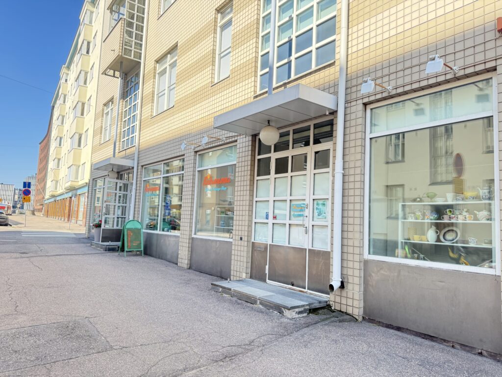 The simple storefront of Retronomi, a Helsinki vintage shop, on a sunny day. The shop is on the ground floor of a cream-colored tiled building, with vintage tableware visible in the display window.