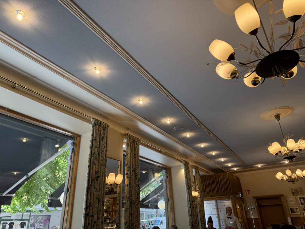 The interior of an elegant restaurant. The bluish-gray ceiling is warmly lit by recessed lights and a chandelier with leaf decorations. Greenery can be seen through the windows, creating a calm atmosphere.