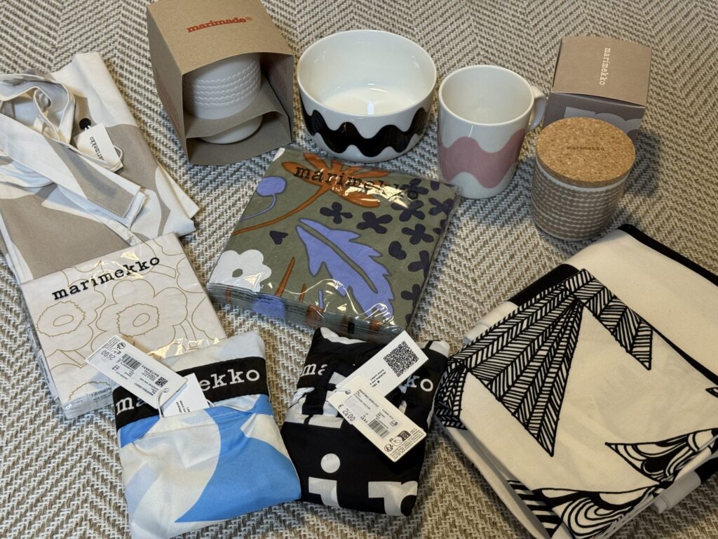 A collection of Marimekko tableware and accessories, including mugs, bowls, reusable bags, and paper napkins, arranged on a herringbone-patterned cloth.