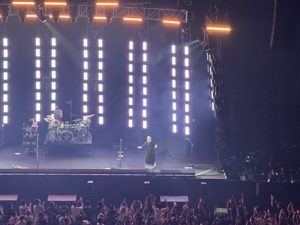 Billy Corgan stands on a concert stage with a drummer and vertical white lights behind him, spreading his arms wide to thank the audience in front of him.