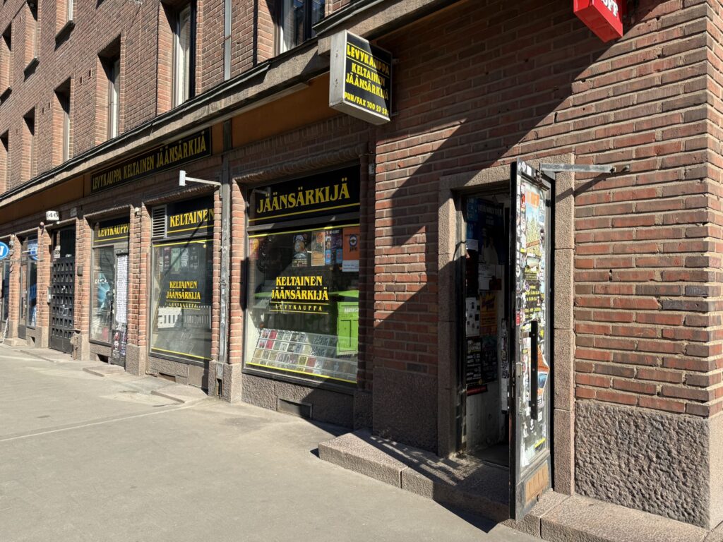 The exterior of the brick record store 'Levykauppa Keltainen Jäänsärkijä' on a sunny day. The shop's door is open, casting a shadow on the sidewalk.