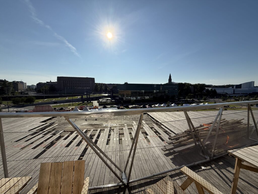 晴れた日に屋上テラスから見下ろしたヘルシンキの街並み。太陽が青空に輝き、手前のウッドデッキの向こうに国会議事堂やOodi図書館などの建物が見える。