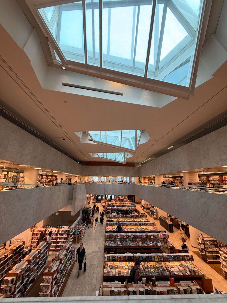 The interior of the Academic Bookstore in Helsinki, viewed from the second floor. The central atrium is illuminated by natural light from large skylights in the ceiling. Bookshelves line the first and second floors, with many people browsing for books.