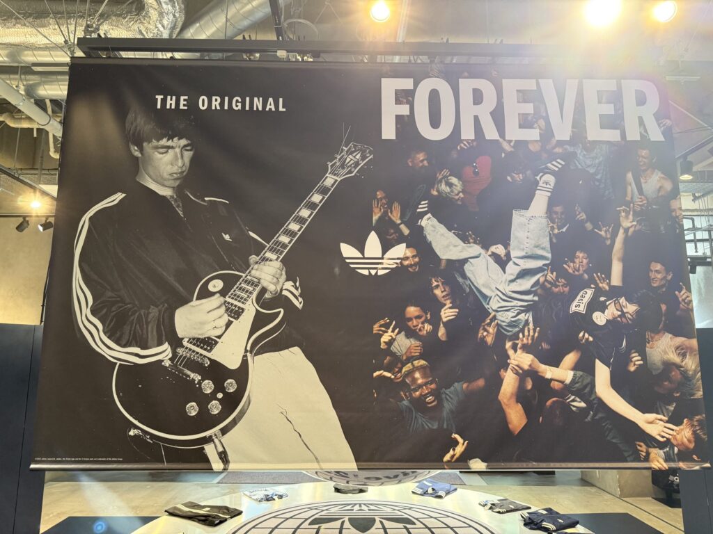 A large Adidas advertising poster on display, featuring a black-and-white image of Noel Gallagher playing guitar and a color image of an energetic concert crowd, with the words 'THE ORIGINAL FOREVER' and the Adidas logo.