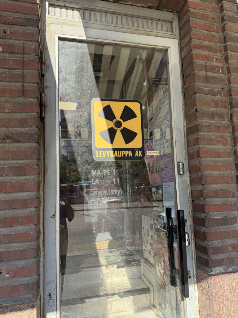 The glass door entrance to the Levykauppa Äx record store, set in a brick building. A yellow and black sticker resembling a radiation symbol serves as the store's logo on the door.