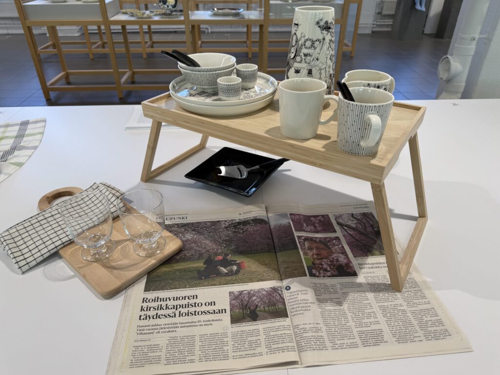木製のベッドトレイの上に、黒い線画模様の食器や花瓶が並べられている展示。手前には新聞とグラスが置かれている。