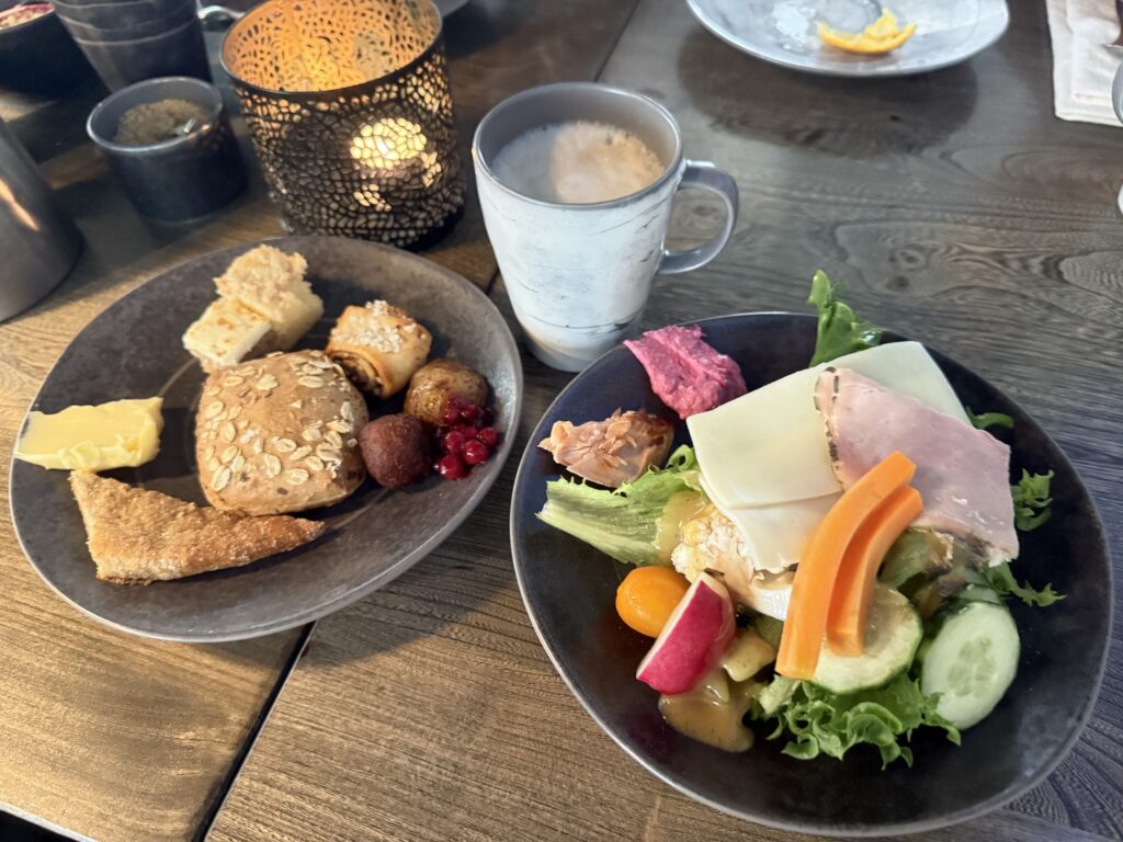 木製のテーブルの上に置かれた2枚の朝食プレートとコーヒー。一方の皿にはパンとバター、もう一方にはサラダ、ハム、チーズが盛られ、背景にはキャンドルの灯りが見える。