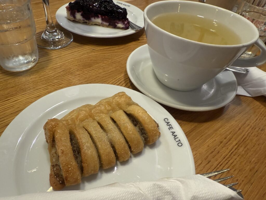 木製のテーブルの上、CAFE AALTOのロゴが入った白い皿に乗ったミートパイと、カップに入ったハーブティー。奥にはブルーベリーチーズケーキも見える。