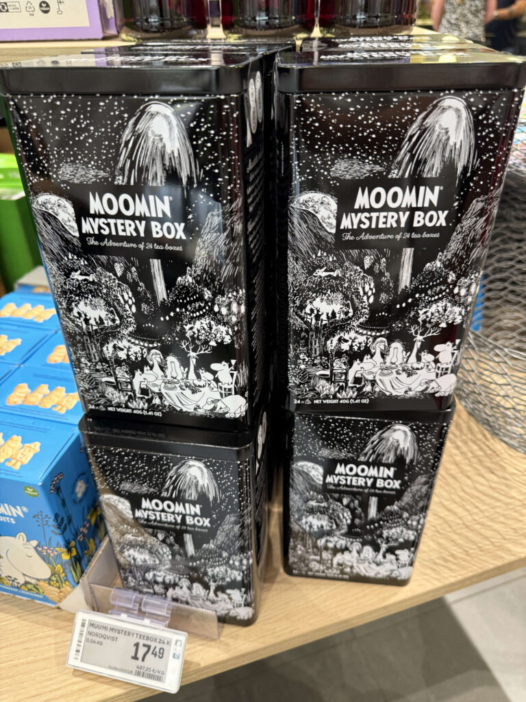 Black tins of 'Moomin Mystery Box' tea stacked on a wooden store shelf, featuring intricate black and white illustrations of Moomin characters in a forest at night.