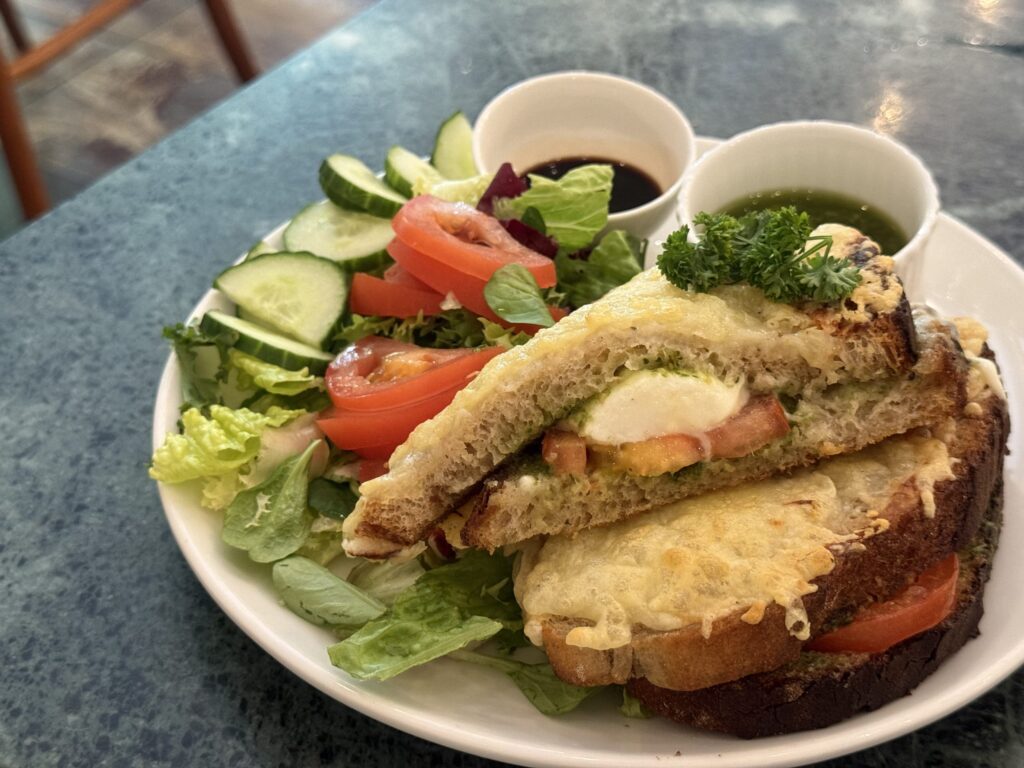 A grilled cheese sandwich cut in half on a white plate, with melted cheese oozing out. It is served with a side salad of tomatoes and cucumbers, and two small cups of dressing.