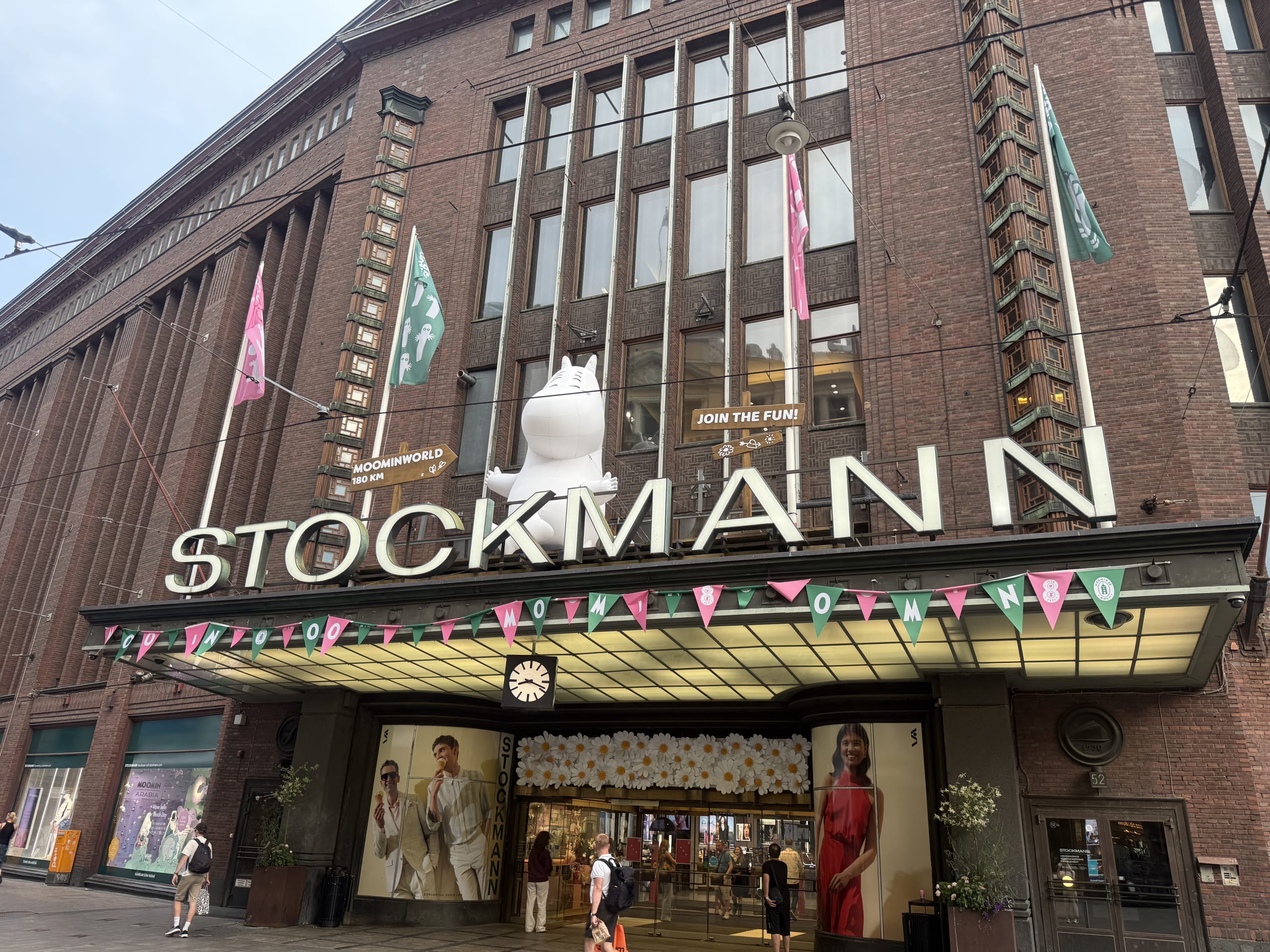 The exterior of the Stockmann department store in Helsinki. A giant, white, inflatable Moomin is sitting behind the large 'STOCKMANN' sign on the brick building's facade.