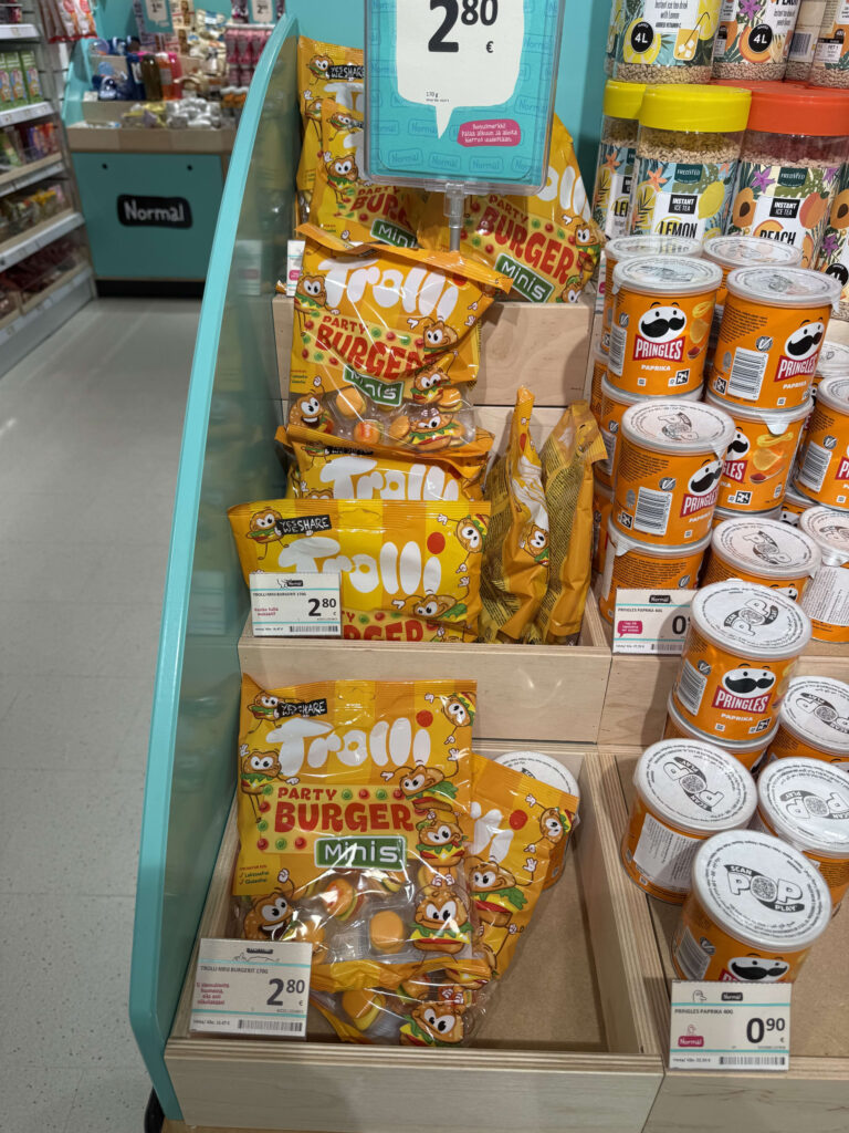 A view of a supermarket shelf displaying bags of Trolli hamburger-shaped gummies and orange cans of Pringles.
