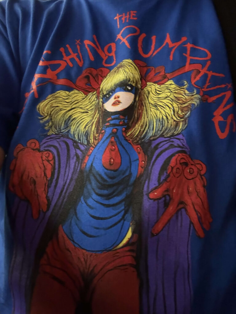 A close-up of a blue The Smashing Pumpkins tour t-shirt, featuring a colorful comic-style illustration of a masked girl with blonde pigtails.