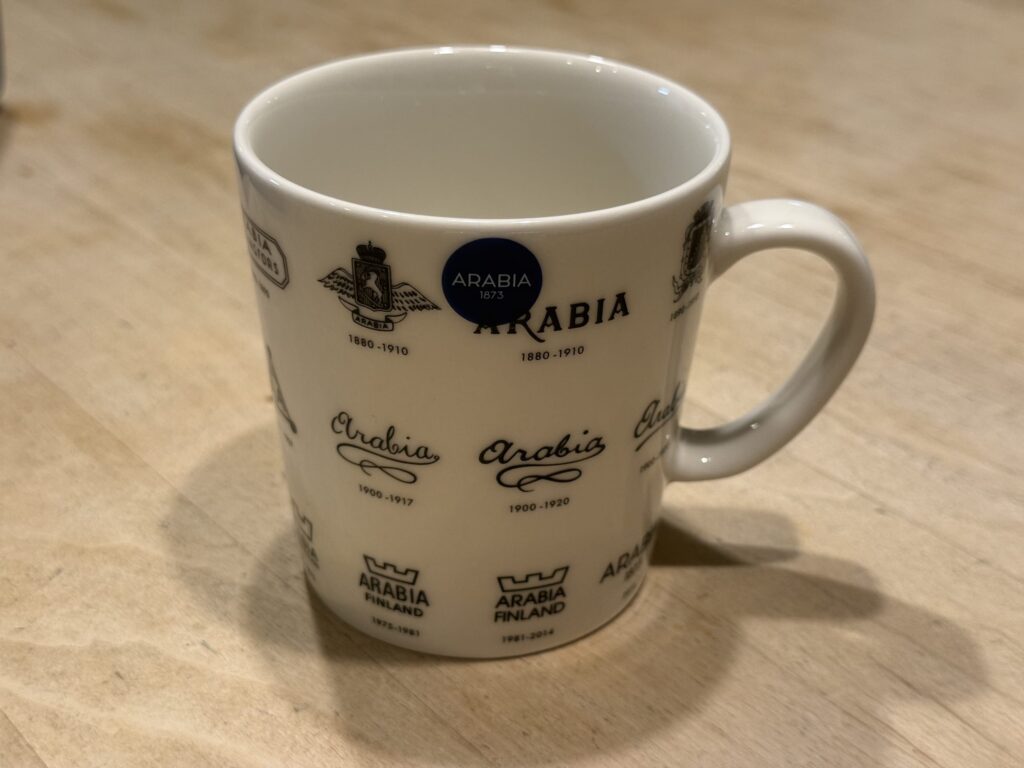 A white mug featuring historical logos of the Finnish brand Arabia, sitting on a wooden table.