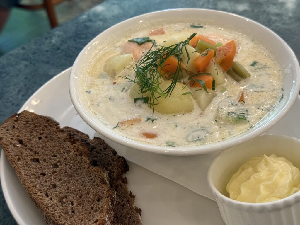 A creamy salmon soup in a white bowl, with visible chunks of potatoes and carrots, garnished with dill. Slices of rye bread and a side of butter are placed next to it.