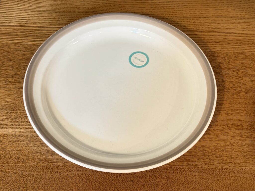 A white circular plate with a beige rim, placed on a wooden table. There is a light blue sticker on the plate that says 'vintage'.